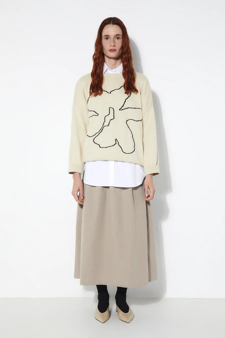 Person wearing a beige Rita Row sweater with a black abstract flower design, white shirt, beige skirt, and black shoes on a white background