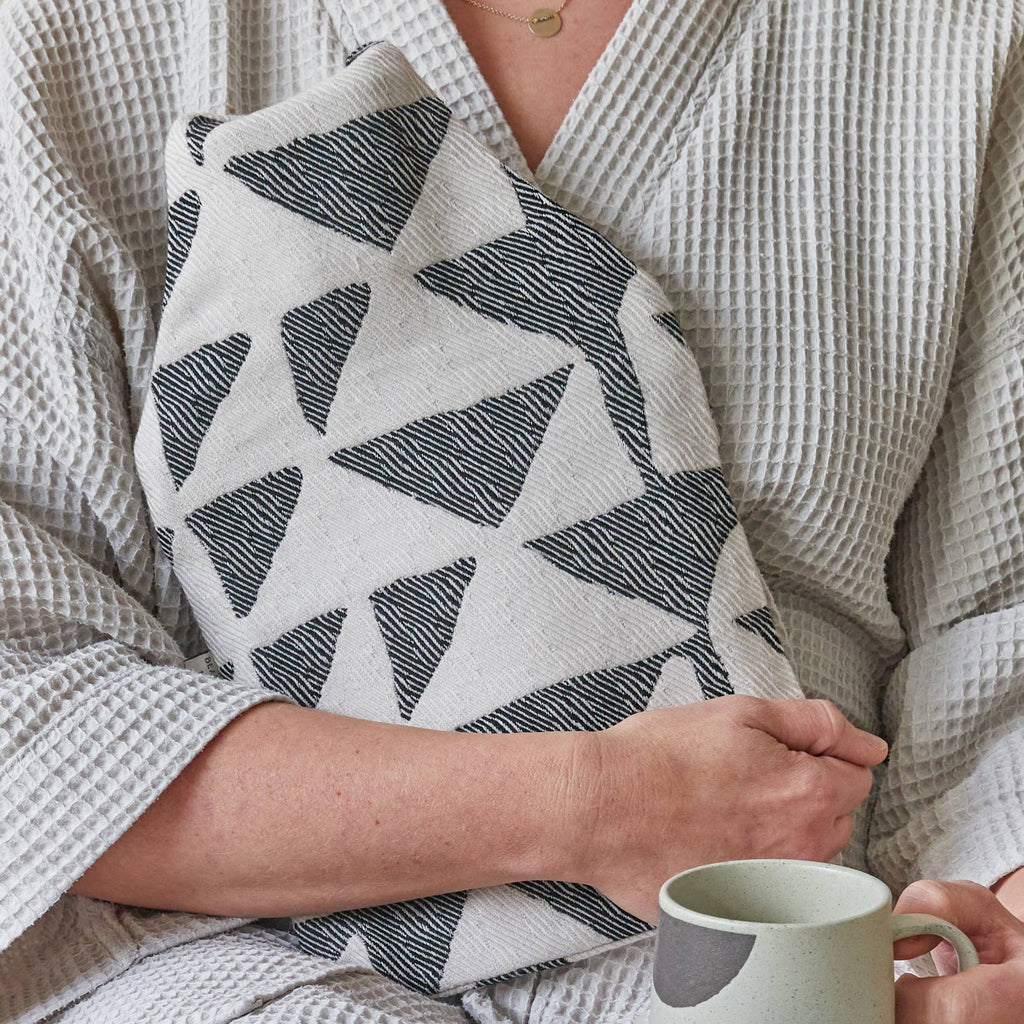 Person holding a Beatrice Larkin Flint Hot water bottle and a mug, wearing a textured robe.