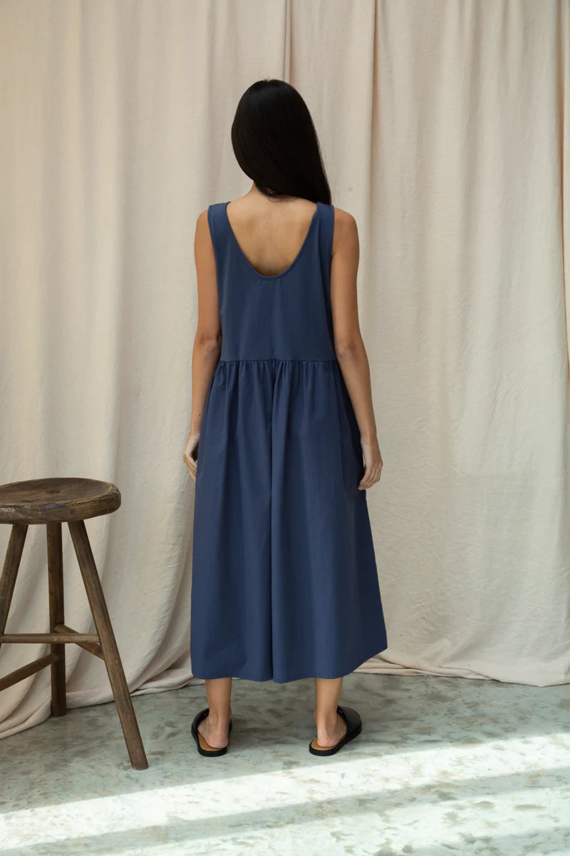 Woman wearing a blue Beaumont Organic  jumpsuit standing in front of a beige curtain.