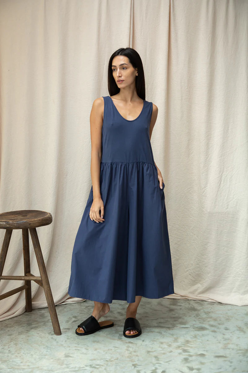 Woman wearing a blue jumpsuit standing in front of a beige curtain.