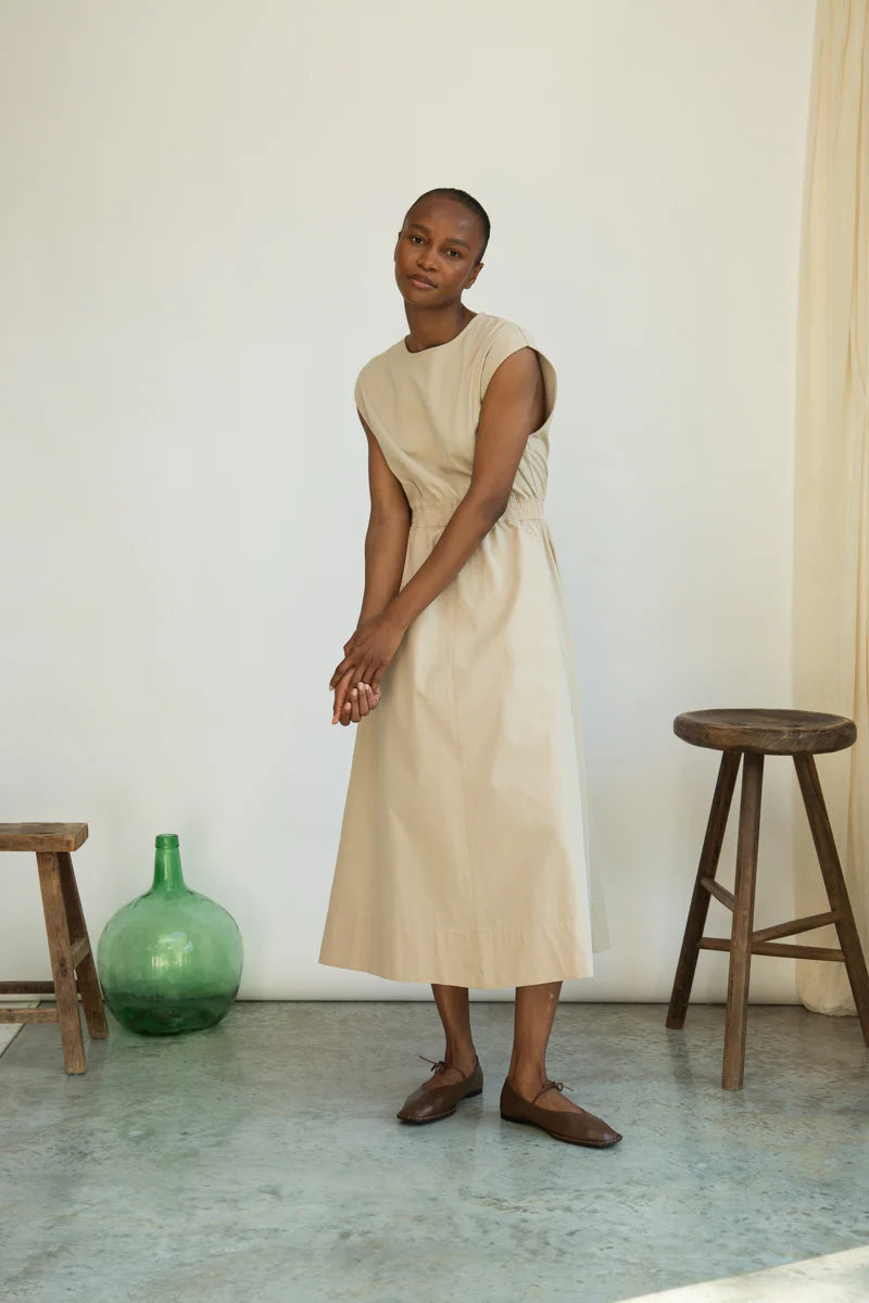Woman in a beige dress standing in a room with wooden stools and a green vase.