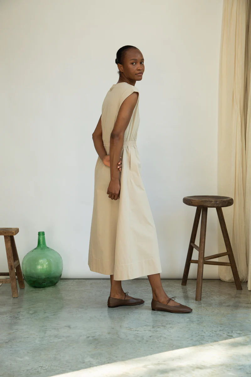 Woman in a beige dress standing in a room with wooden stools and a green vase.