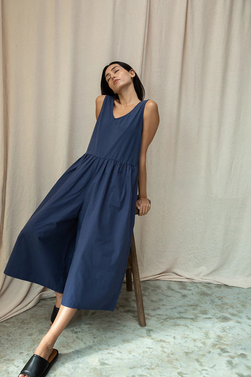 Woman wearing a blue Beaumont Organic jumpsuit  standing against a beige curtain.