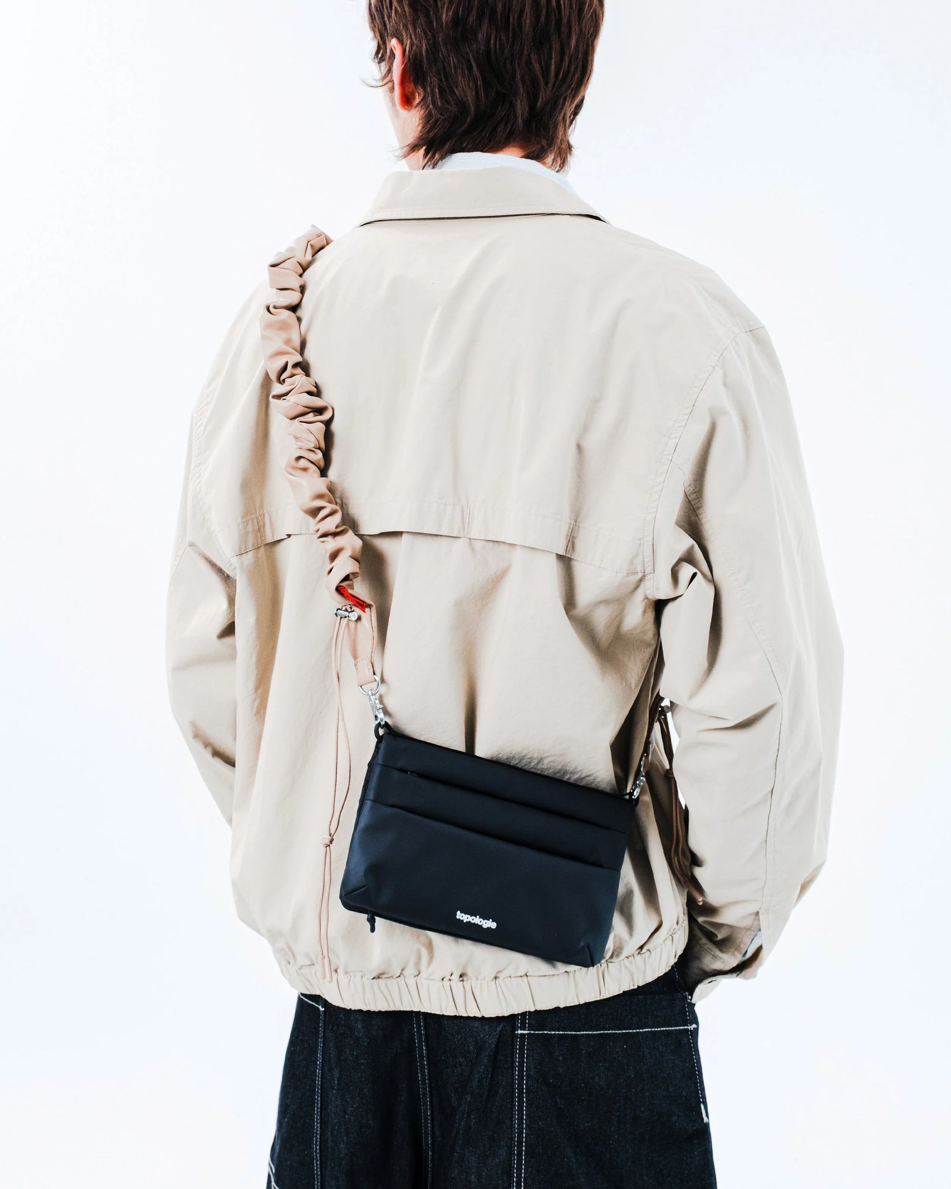 Person wearing a beige jacket with a black crossbody bag on a white background