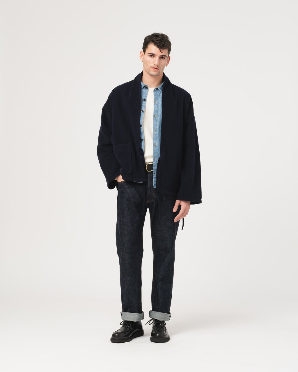 A male model stands against a light gray studio background wearing a dark navy jacket over a light blue and white striped shirt, dark jeans with cuffed hems, and black shoes.