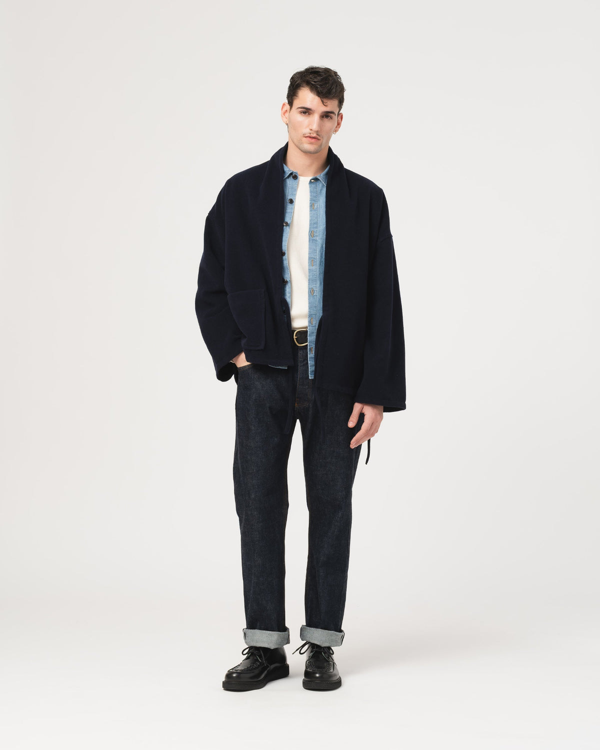 A male model stands against a light gray studio background wearing a dark navy jacket over a light blue and white striped shirt, dark jeans with cuffed hems, and black shoes.