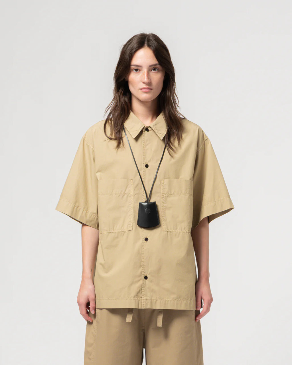 Person wearing a beige short-sleeve girls of dust Eat dust unisex shirt and pants with a black pendant necklace on a white backgrounded 