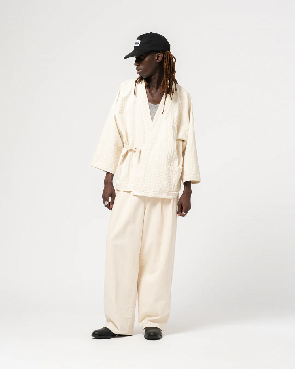 Person wearing a cream-colored outfit with a black cap on a white background