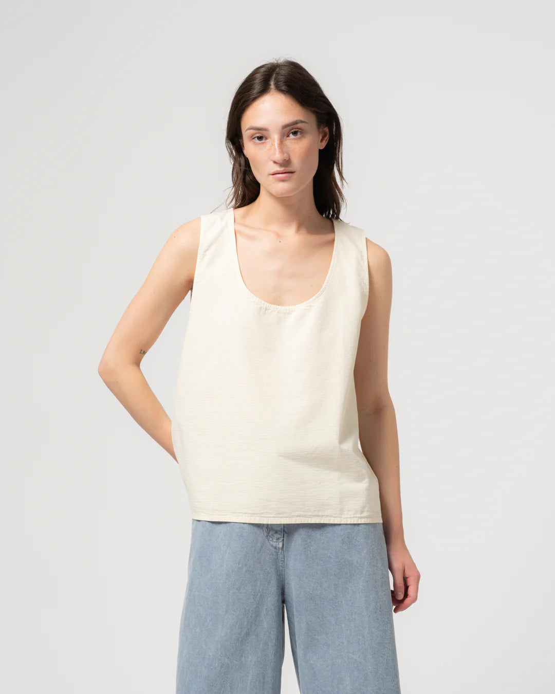 Woman wearing an off white sleeveless top and light blue pants on a plain background