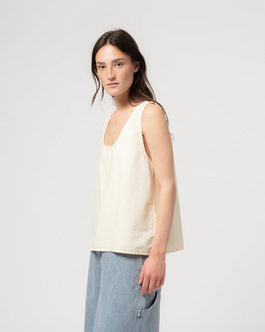 Woman wearing a beige sleeveless top and light blue jeans against a plain background