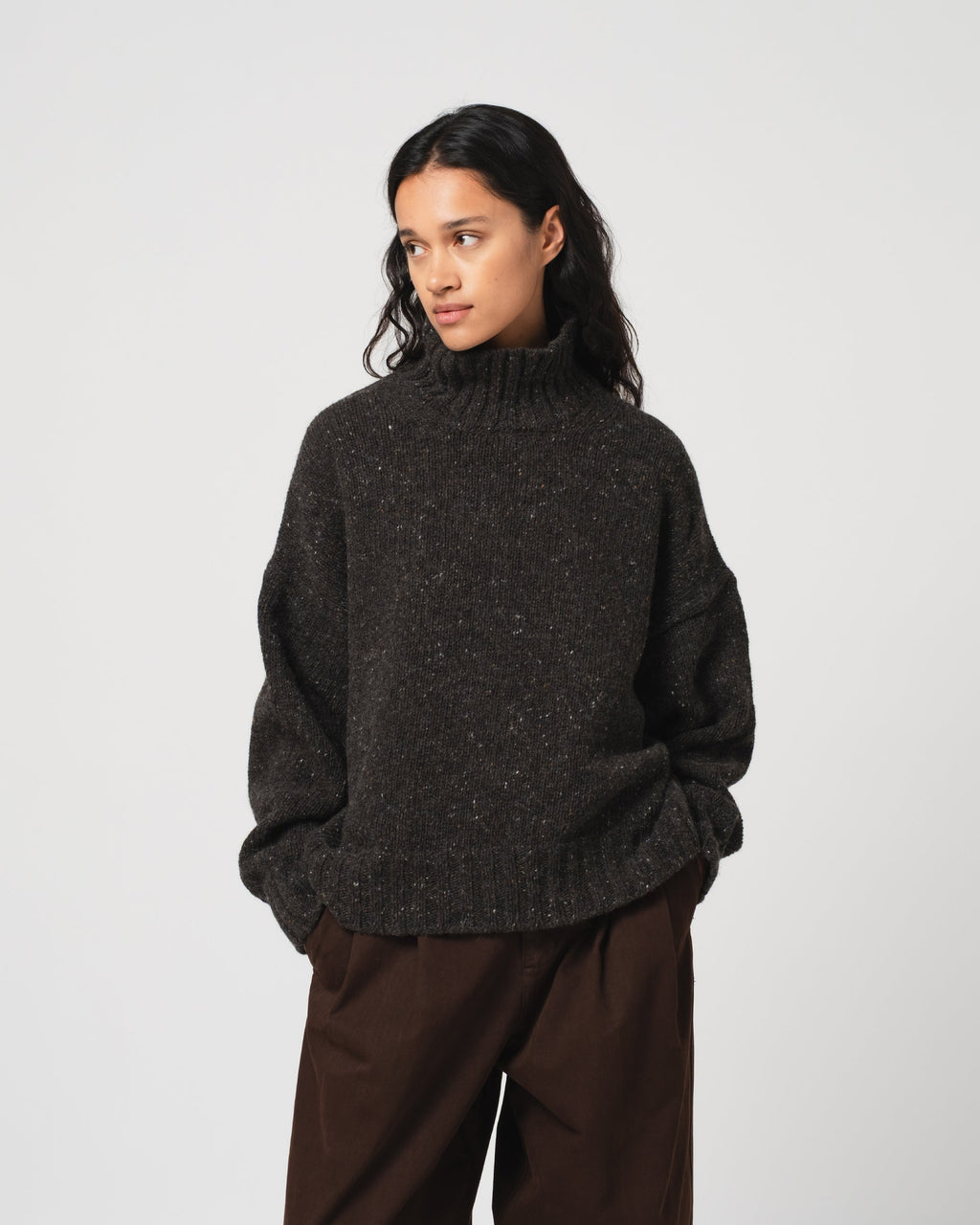 A woman wearing a dark charcoal grey knit mock neck sweater and dark brown trousers, standing against a light gray studio background.