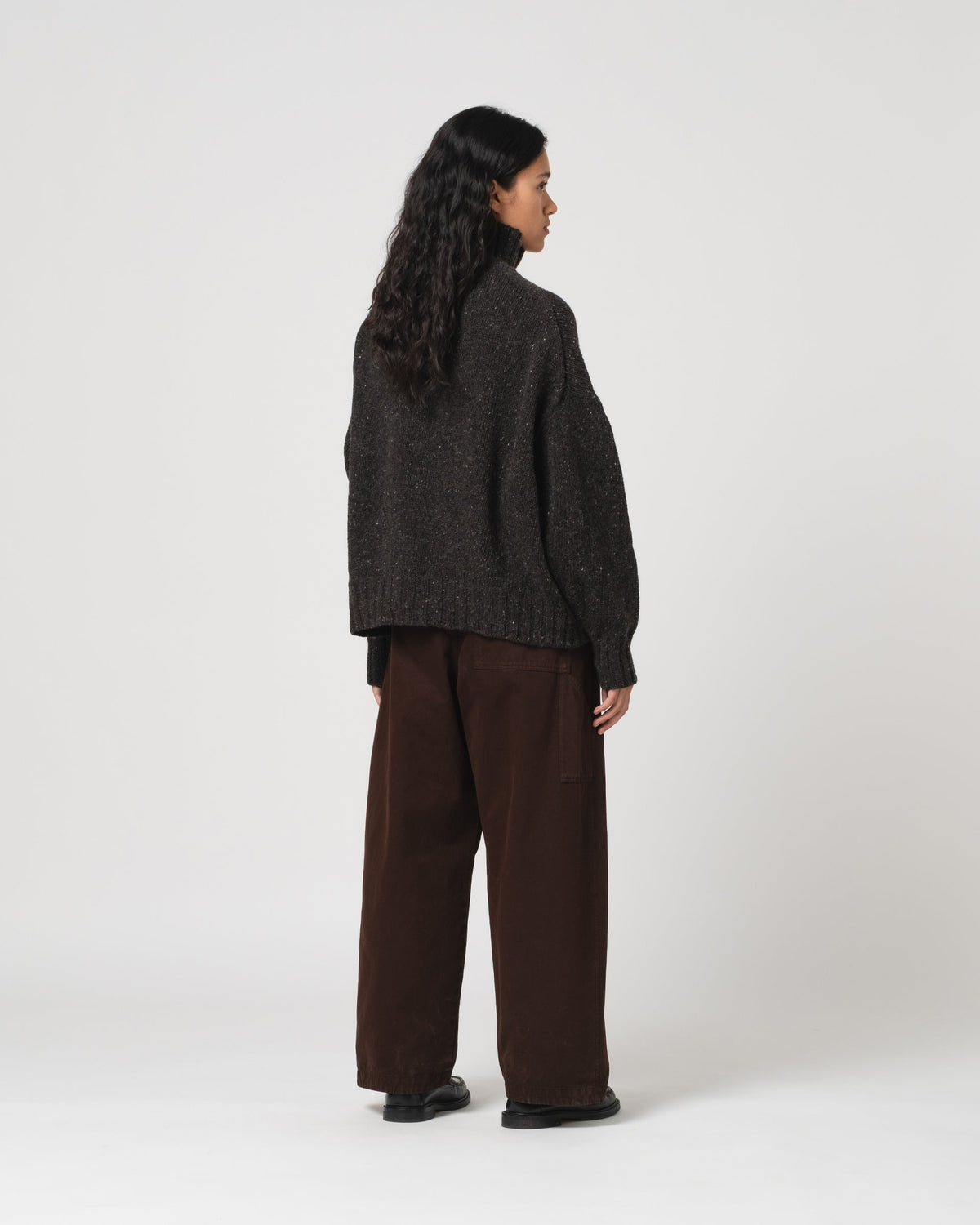 A woman is shown from the back wearing a dark charcoal grey Girls of Dust knit sweater and dark brown wide-leg trousers, standing against a plain light grey studio background.