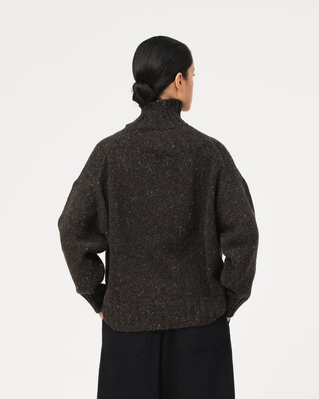 A model shown from the back wearing a dark grey Girls of Dust Cornish Sweater Neppy Wool Asphalt  textured knit turtleneck sweater and black wide-leg trousers against a light grey  studio background.