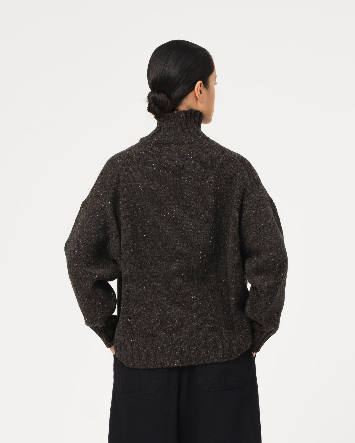 A model shown from the back wearing a dark grey Girls of Dust Cornish Sweater Neppy Wool Asphalt  textured knit turtleneck sweater and black wide-leg trousers against a light grey  studio background.