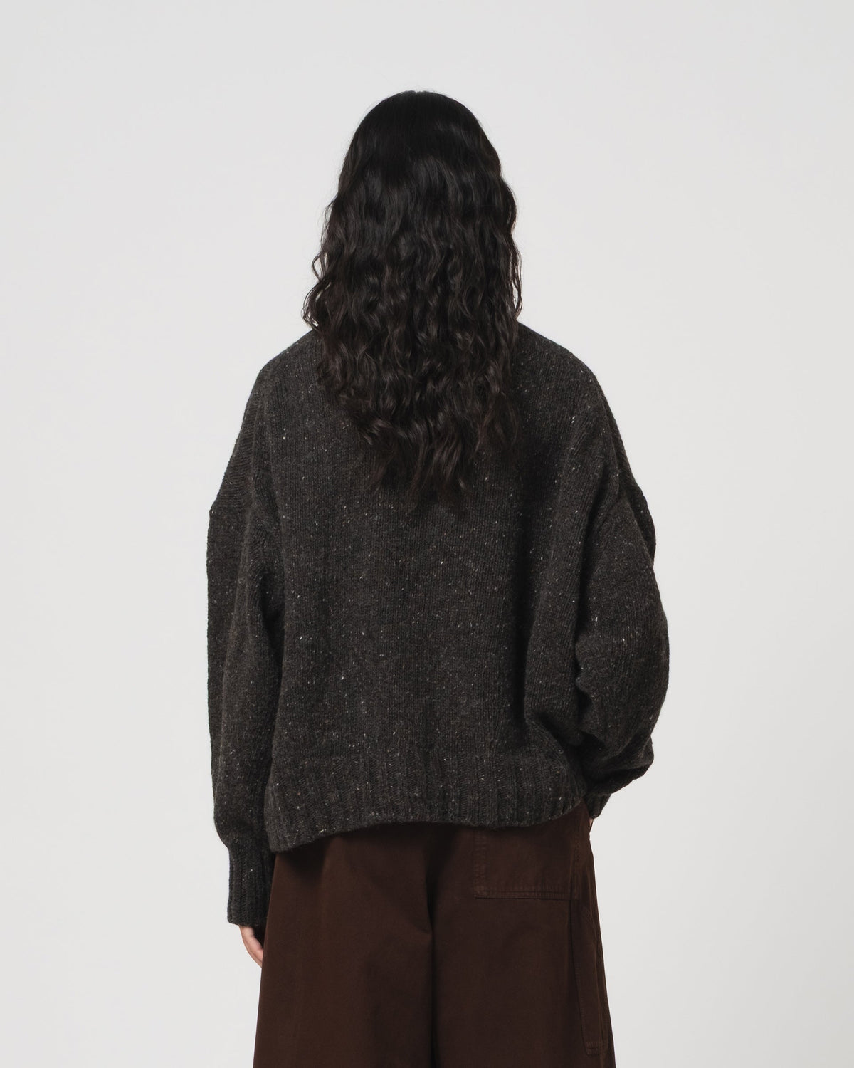 A person shown from the back wearing a dark charcoal grey knit sweater with subtle flecks and brown wide-leg pants, photographed against a plain light gray studio background.