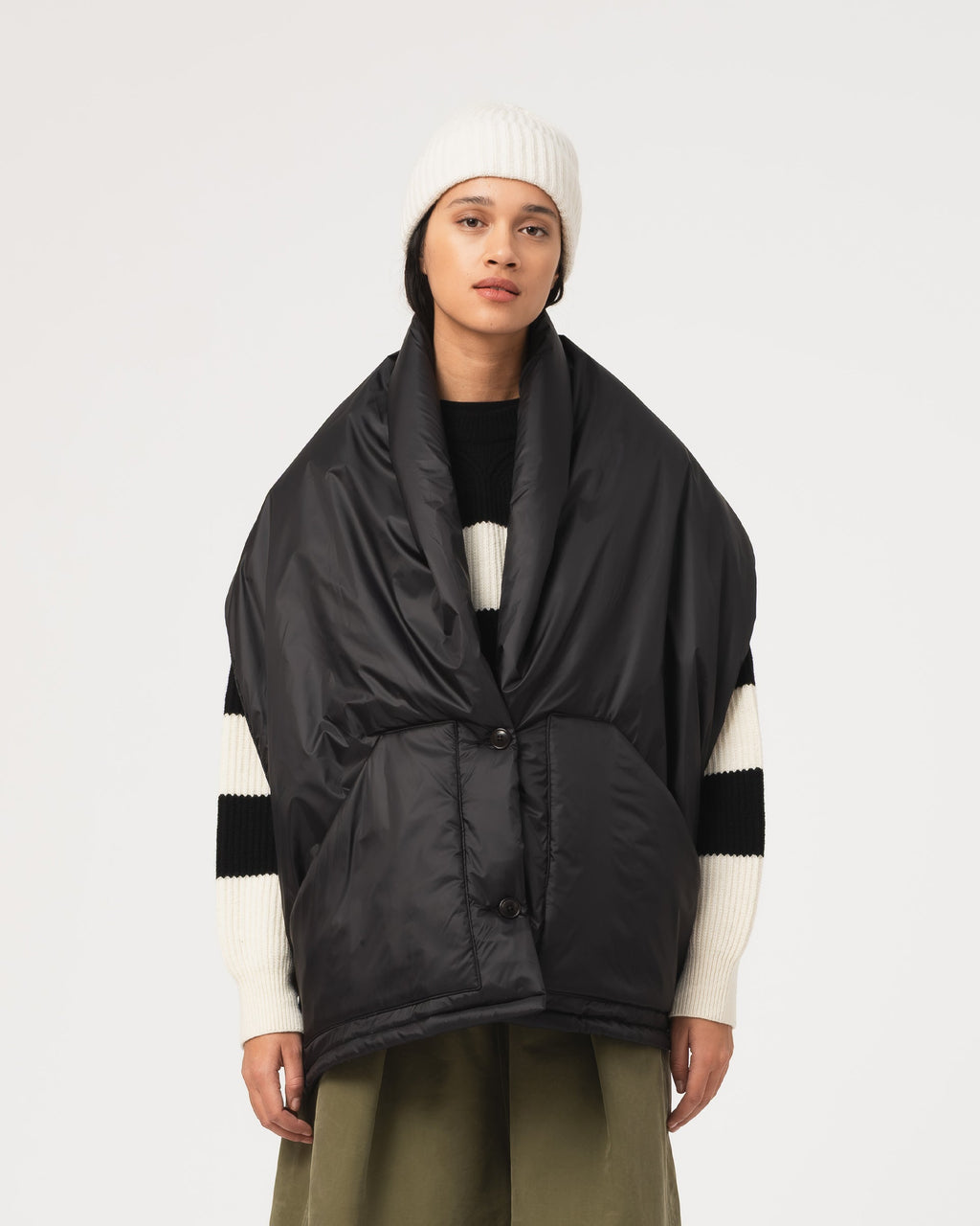 A model wearing a black oversized puffer wrap jacket with white striped knit sleeves, a white beanie, and olive green pants, photographed against a light gray studio background.