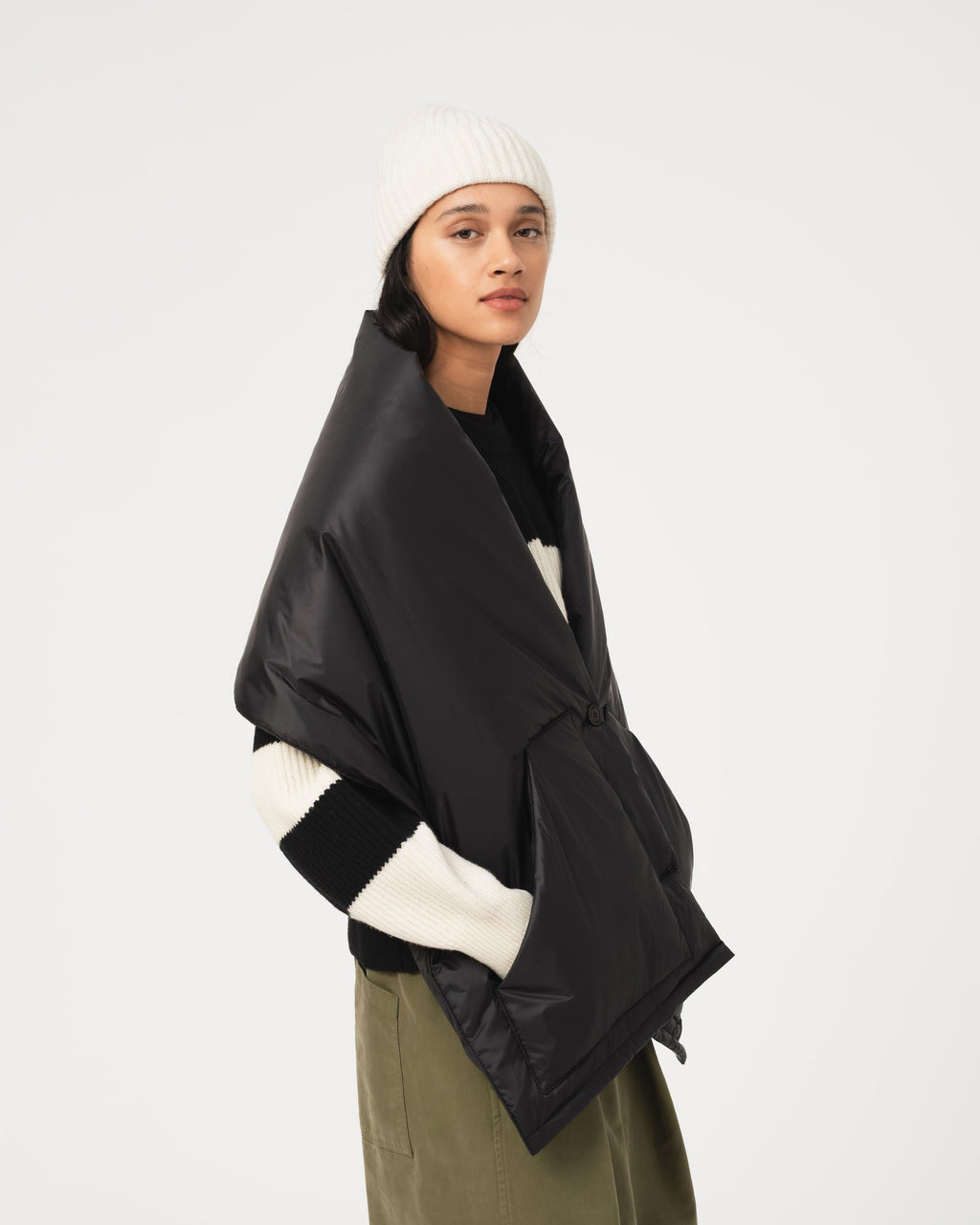 A model wearing the Girls of Dust black puffer scarf over a black and white knit sweater, olive green pants, and a white beanie, photographed against a solid light gray background.