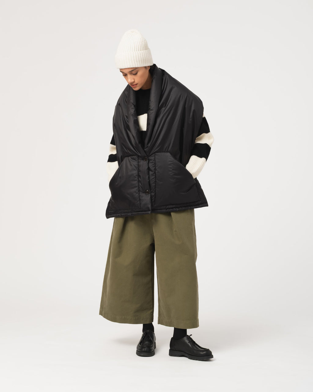 A model wearing Girls of Dust  black quilted puffer scarf with cream sleeve stripes, olive green wide-leg pants, a white knit beanie, and black shoes, photographed against a plain light gray studio background.