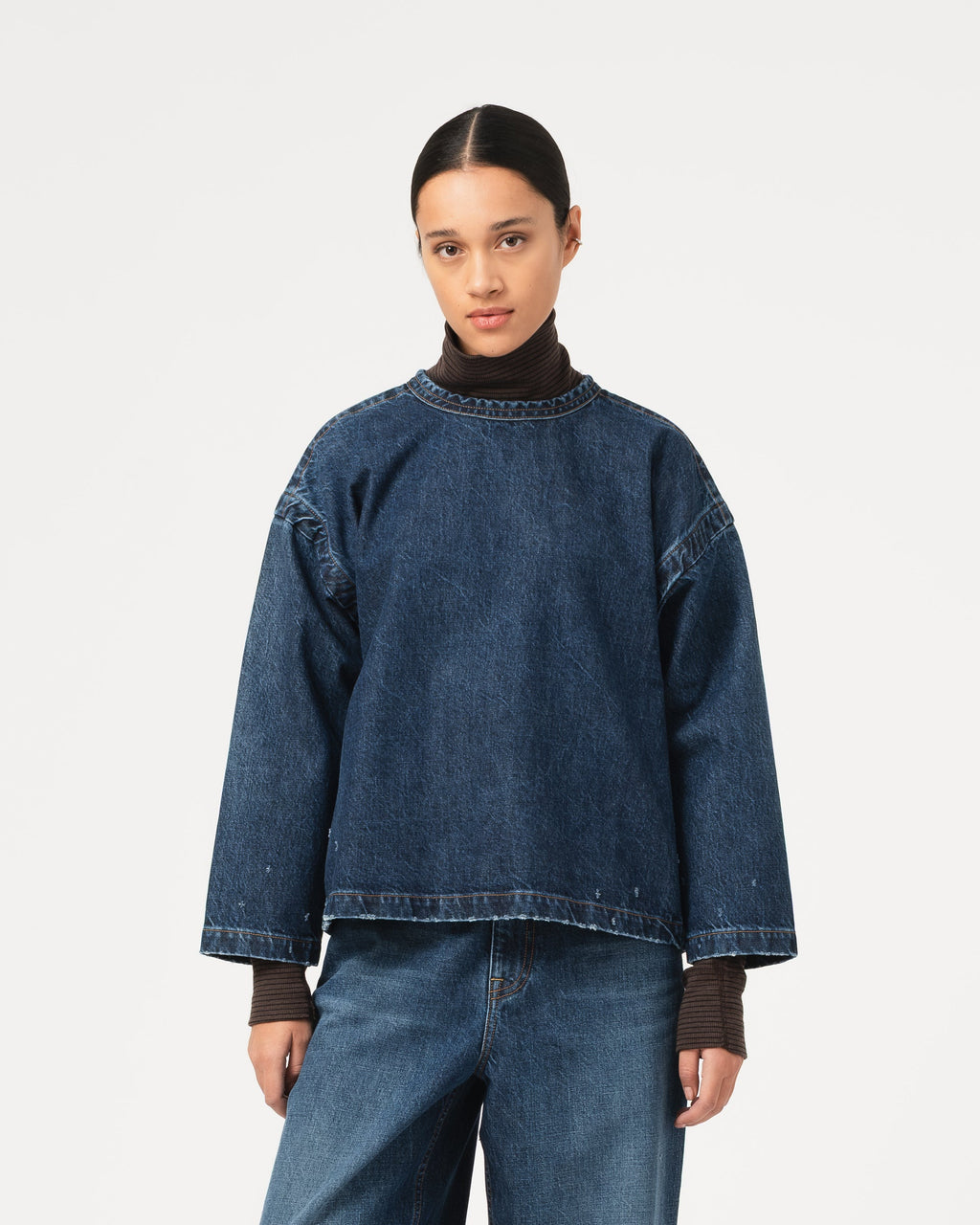 A model wearing a dark blue cropped denim top with a crew neck, layered over a dark brown turtleneck sweater and paired with wide-leg blue jeans, set against a plain light gray background.
