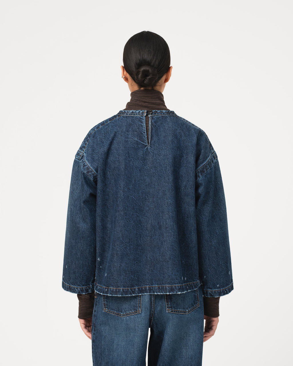 A model viewed from the back wearing a dark blue denim Girls of Dust Guard Smock Century smock  top over a dark brown turtleneck and matching blue jeans, set against a plain light grey studio background.