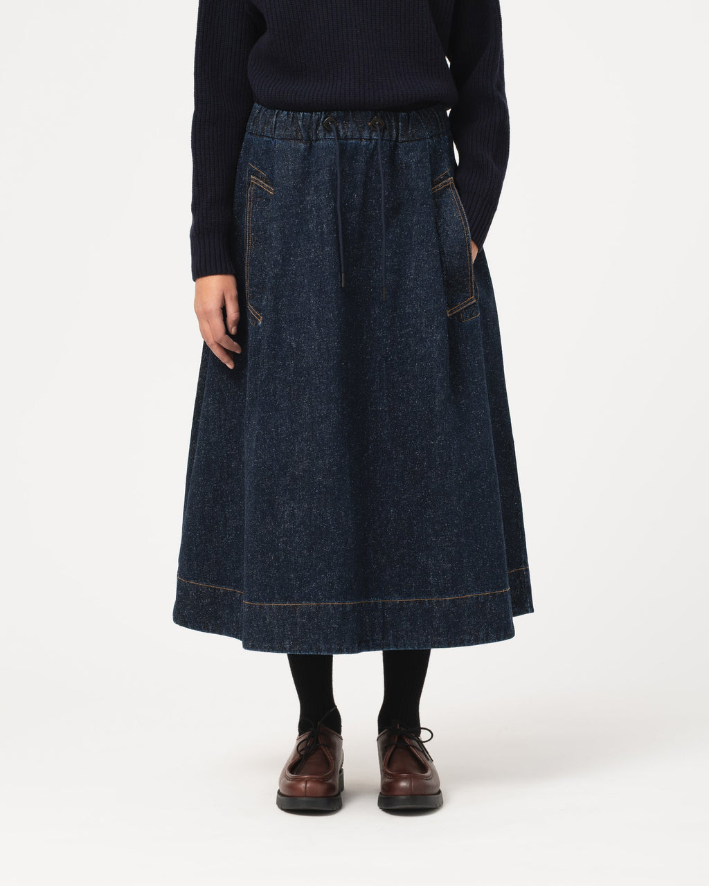 A person wearing a dark blue denim midi skirt with front patch pockets, paired with a dark sweater, black socks, and brown leather shoes, photographed against a plain light gray studio background.
