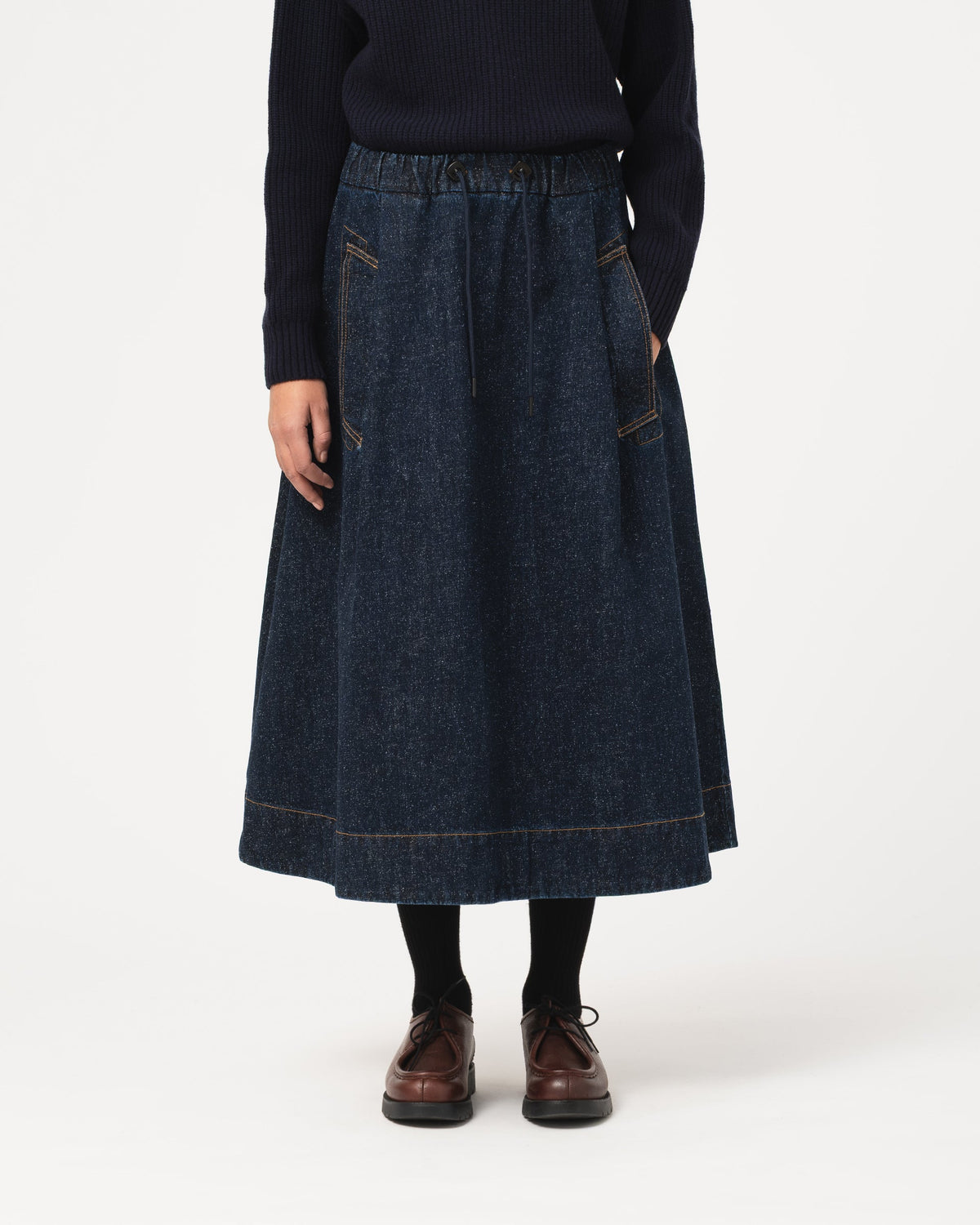 A person wearing a dark blue denim midi skirt with front patch pockets, paired with a dark sweater, black socks, and brown leather shoes, photographed against a plain light gray studio background.