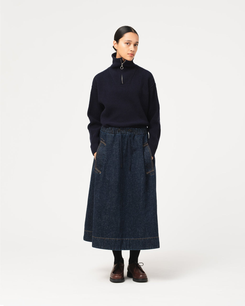 A model stands against a light gray studio background wearing a dark navy mock-neck quarter-zip sweater and a dark blue denim Girls of Dust Meadow Skirt Neppy Denim Indigo midi skirt with  pockets.