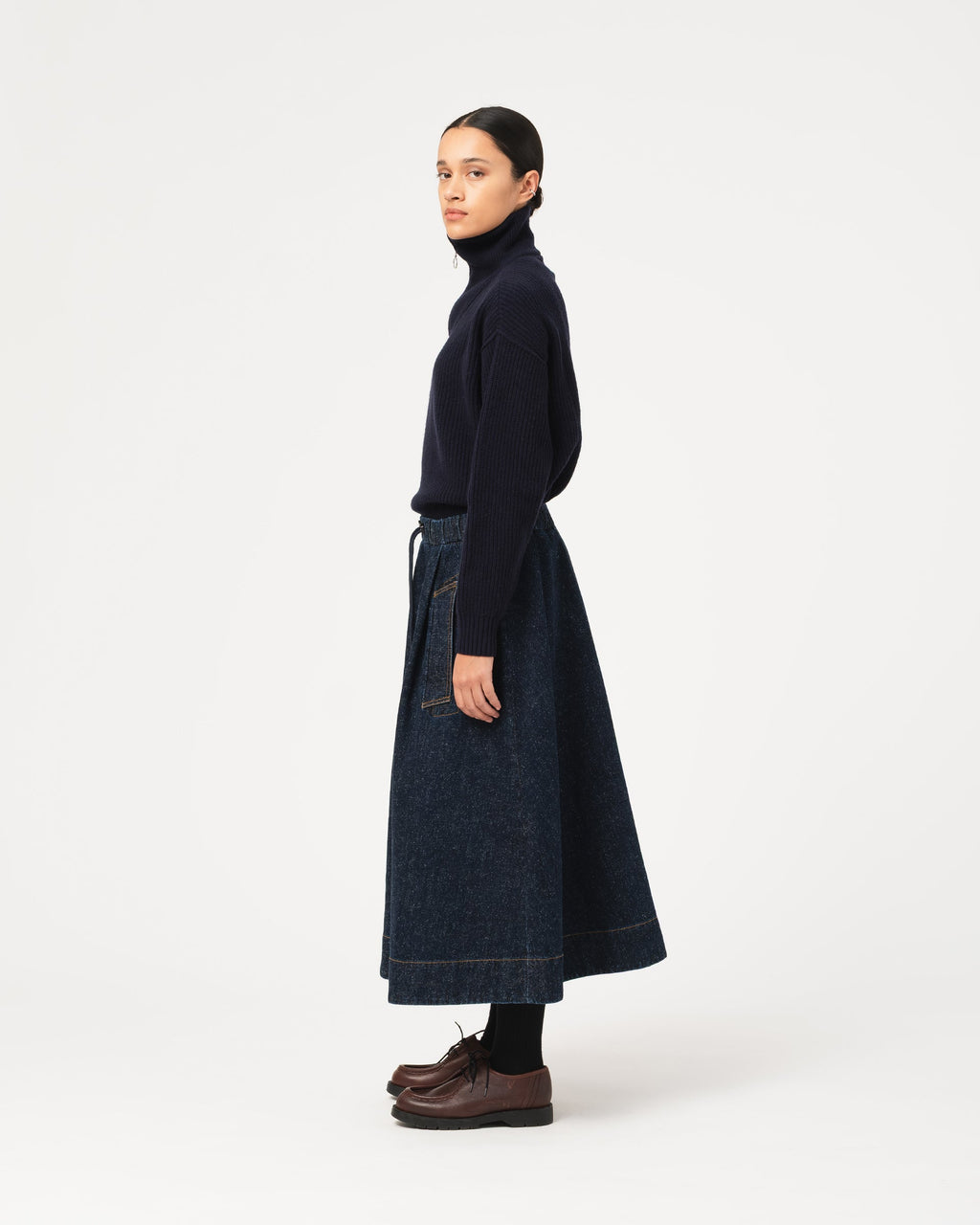 A model is shown in profile wearing a dark navy blue turtleneck sweater and Girls of Dust Meadow Skirt Neppy Denim Indigo  dark blue denim midi skirt with a tie waist, paired with dark brown leather shoes against a plain white studio background.