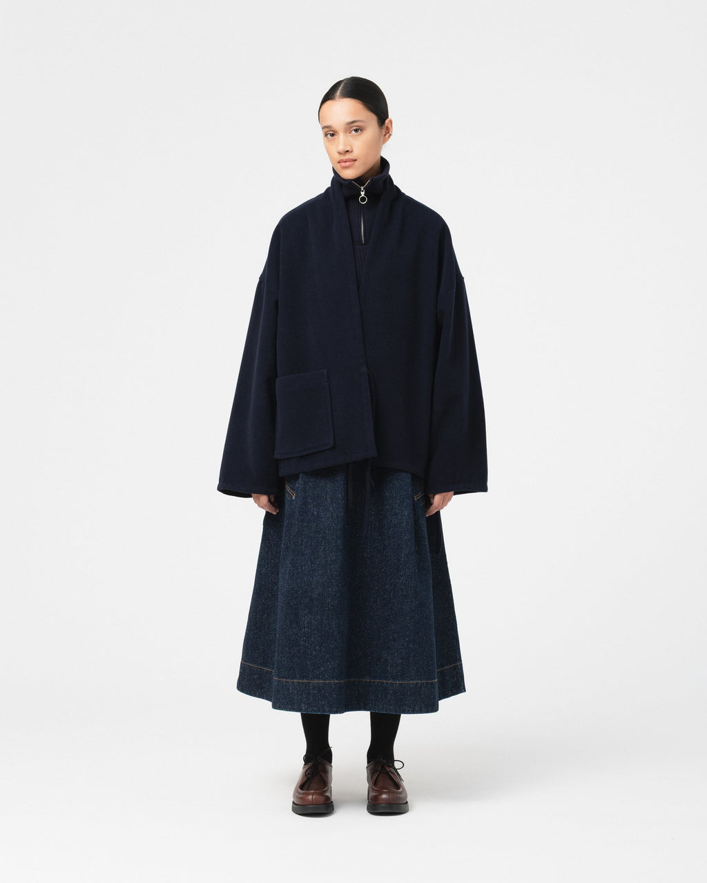 A model stands against a light gray studio background wearing a Girls of Dust Meiji Kimono in navy wool  with a high collar and front patch pocket over a dark blue denim skirt, paired with black socks and brown shoes.