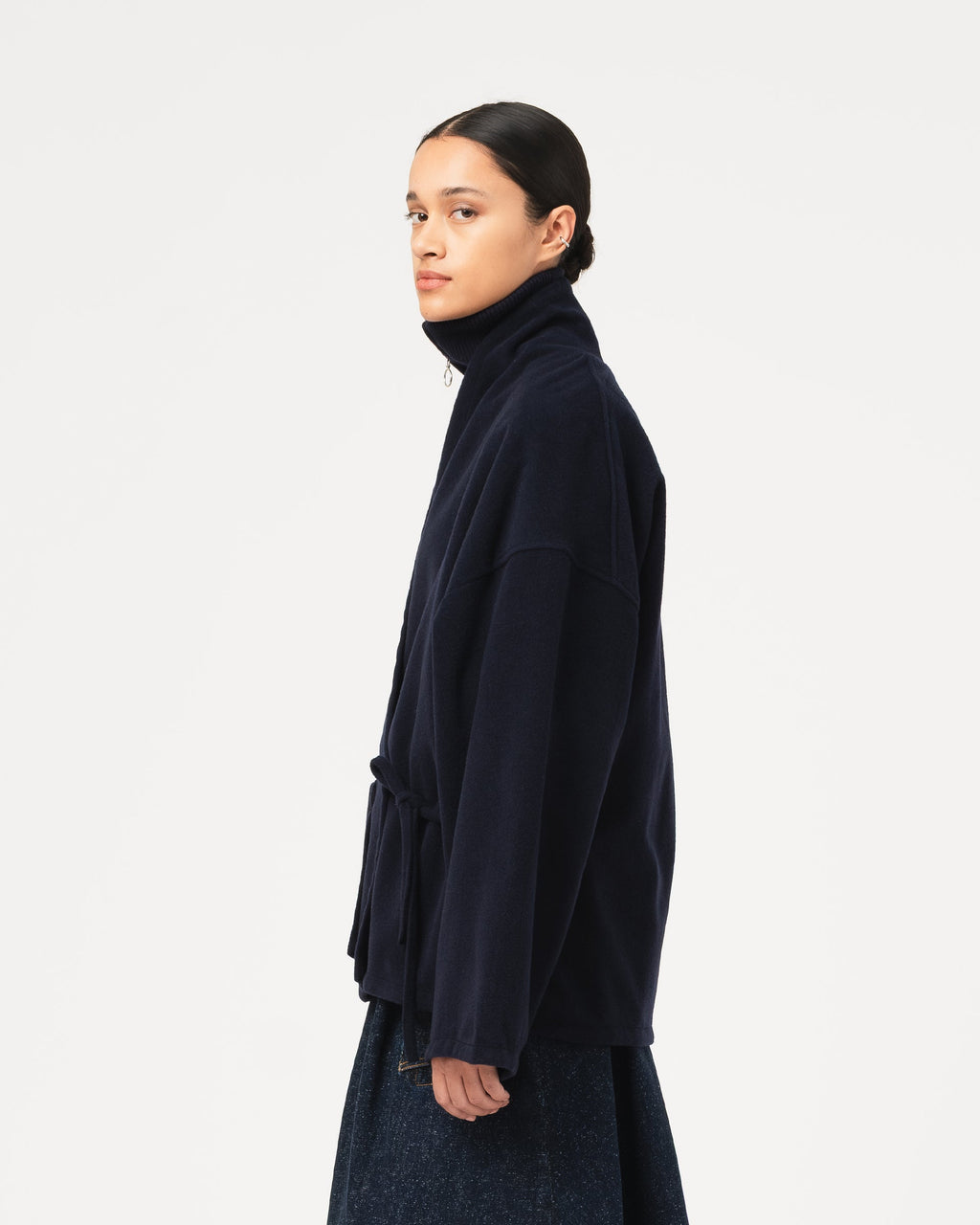 A model shown in profile wearing a dark navy blue Girls of Dust Meiji Kimono in Pachuca Wool with a dark denim skirt against a plain light gray studio background.