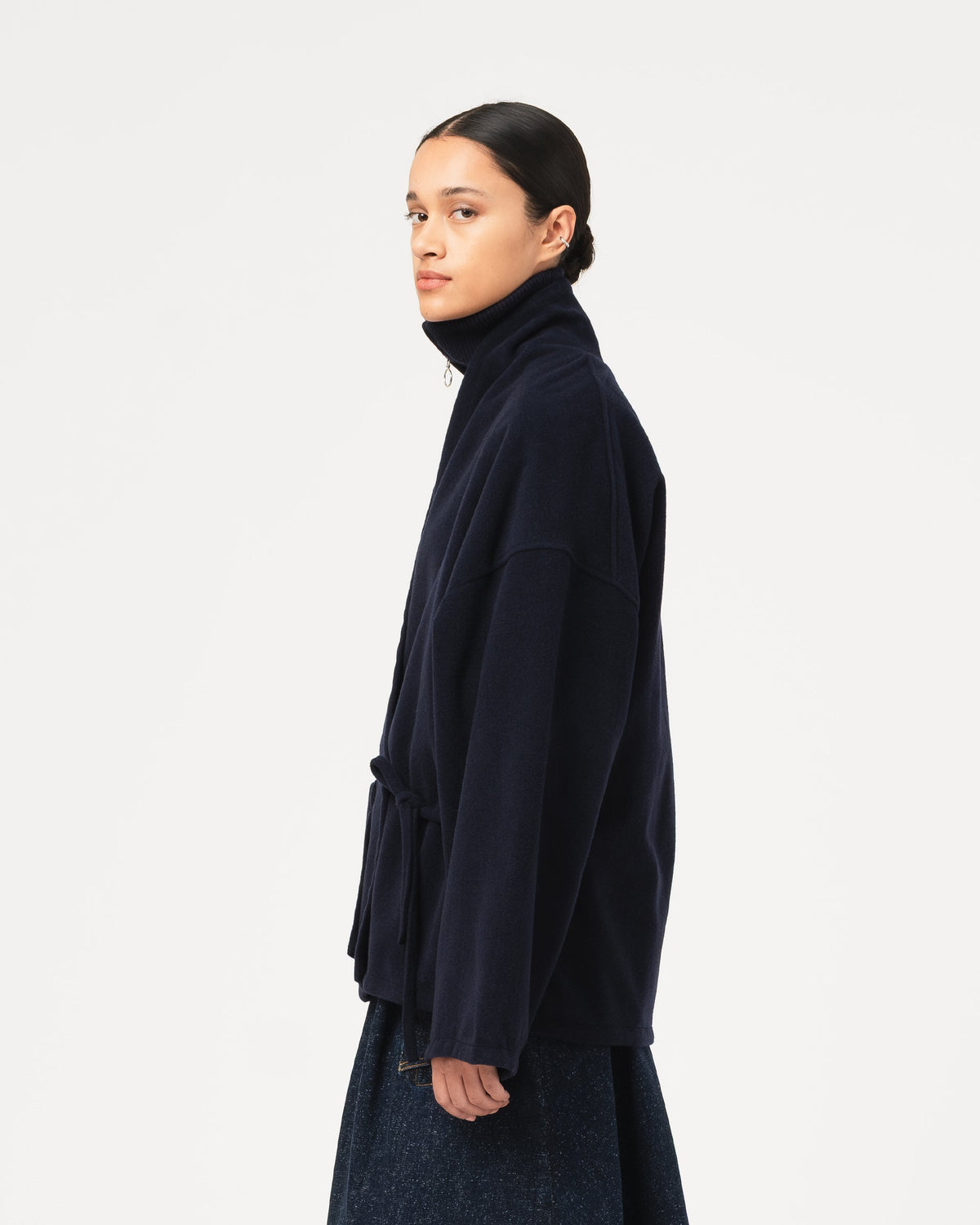 A model shown in profile wearing a dark navy blue Girls of Dust Meiji Kimono in Pachuca Wool with a dark denim skirt against a plain light gray studio background.