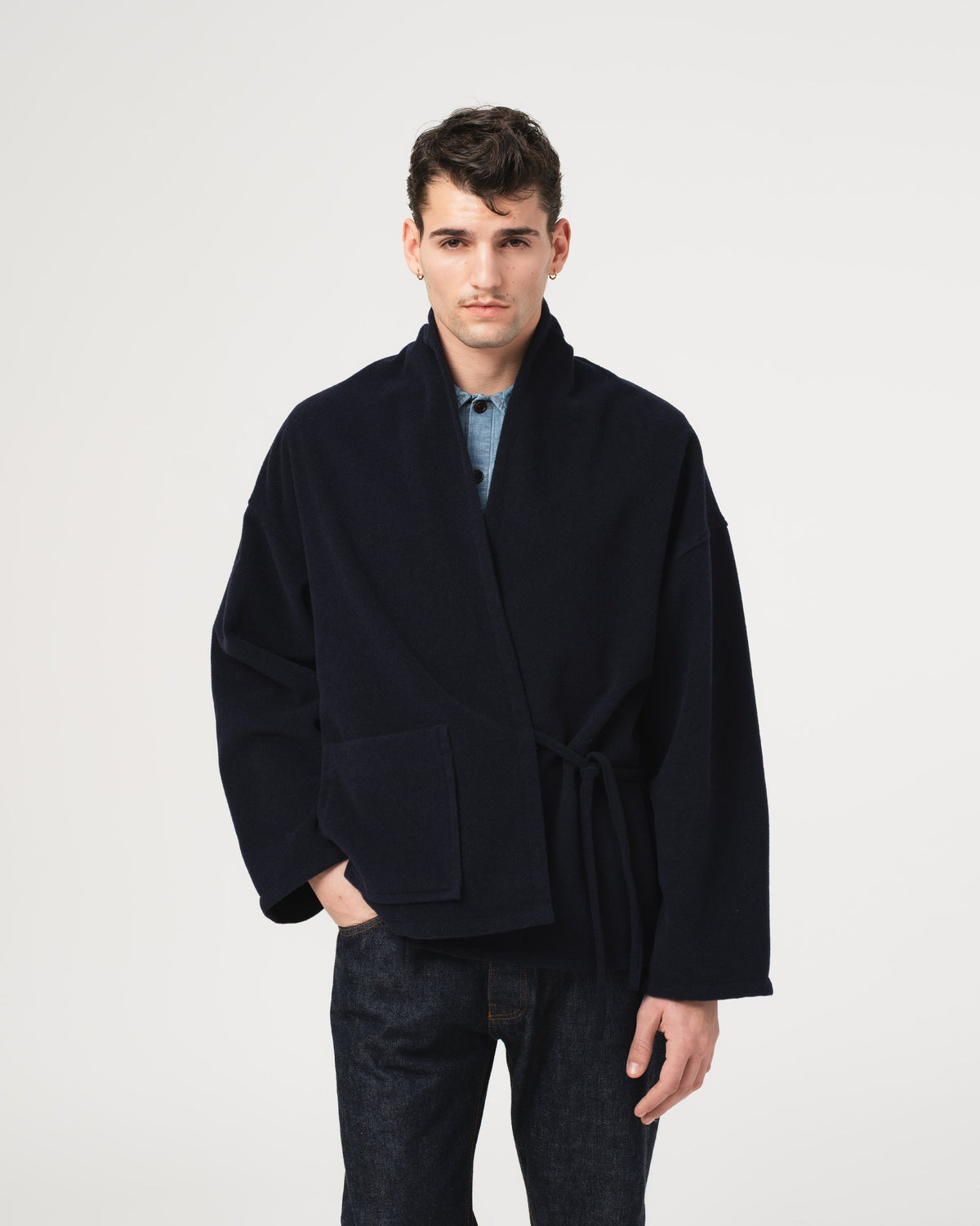 A man wearing a dark navy blue Eat Dust Meiji Kimono Pachuca Wool Navy jacket with a tie belt, styled over a light blue shirt and dark jeans, standing against a plain light gray background.
