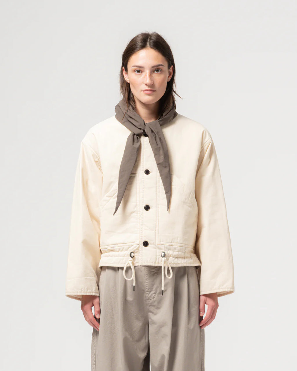 Person wearing a cream jacket with black buttons and beige pants, standing against a plain background.