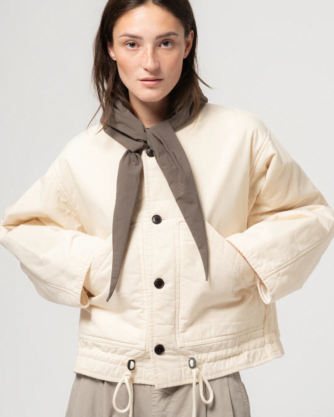 Woman wearing a Ecru Girls of Dust Neutron Jacket quilted jacket with a brown scarf against a white background