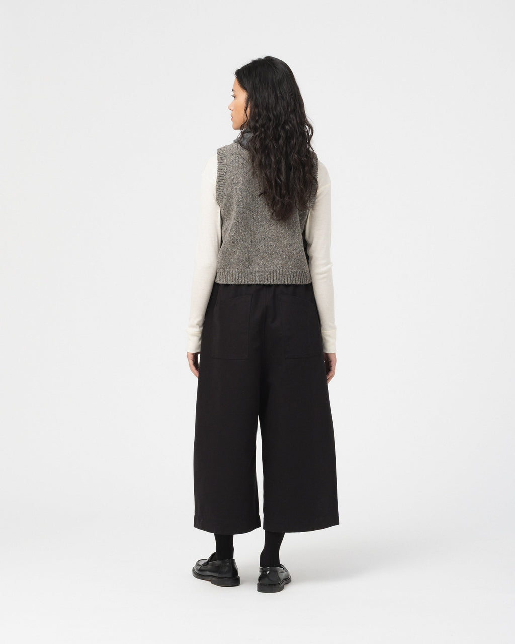 A model shown from the back wearing a grey knit sleeveless vest over a cream long-sleeve top, paired with wide-leg black trousers and black shoes, against a light grey studio background.