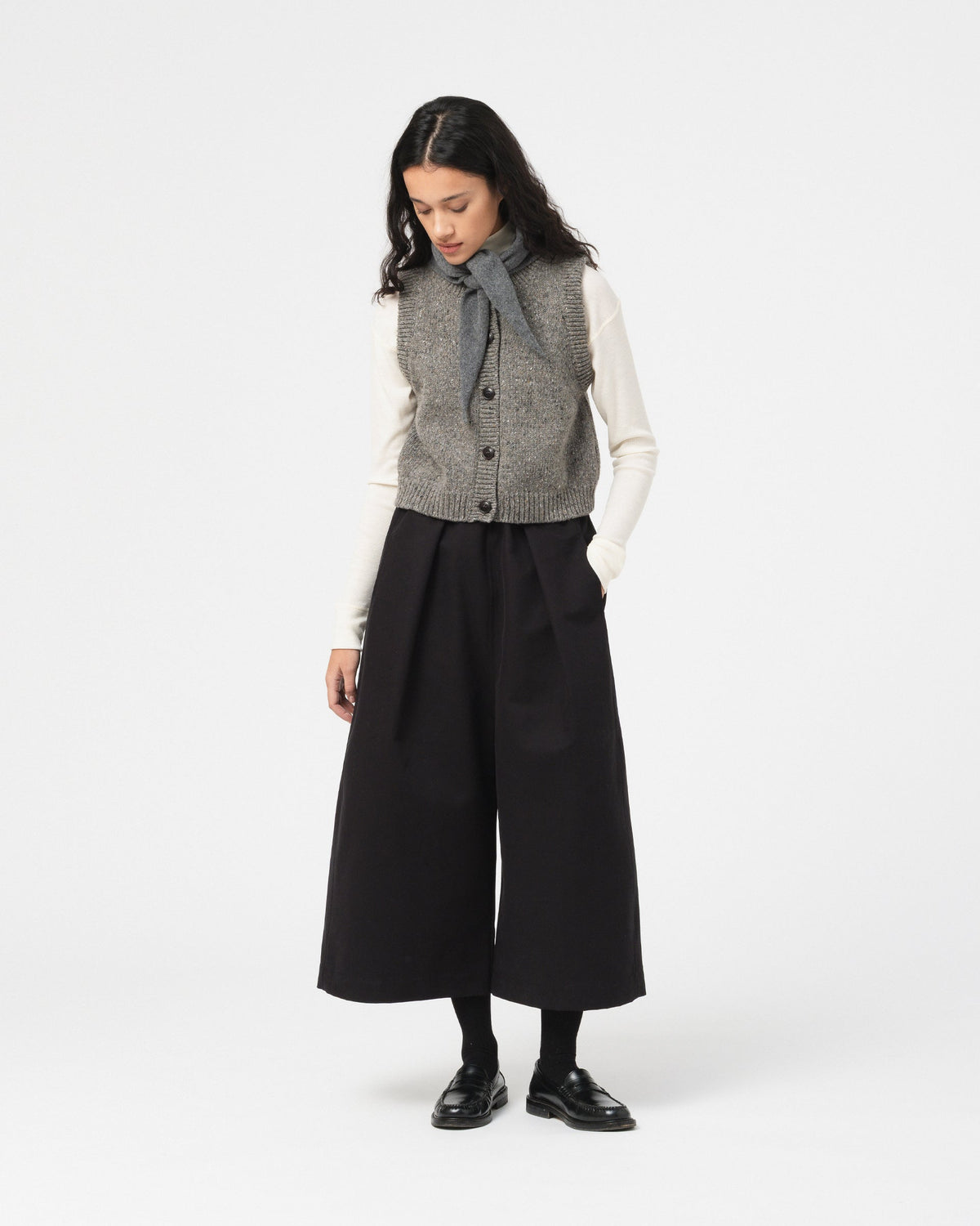 A model wearing a Girls of Dust grey textured wool vest over a white long-sleeve top, a grey tie-neck scarf, black wide-leg cropped pants, black tights, and black leather shoes, photographed against a solid light gray studio background.