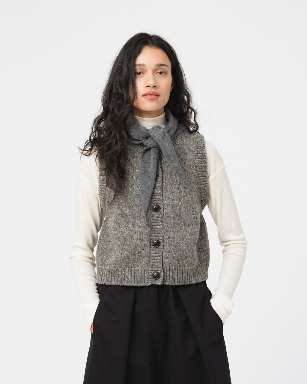 A model wearing a Girls of Dust grey knit button-front vest over a cream turtleneck sweater and a matching grey knotted scarf, paired with a black trousers, against a plain light grey studio background