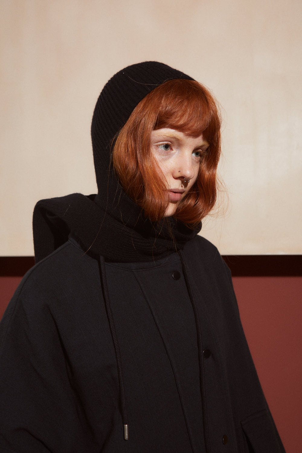 A model with red hair wearing a black knit hooded beanie and a black button-front coat, photographed against a beige and dark red background.