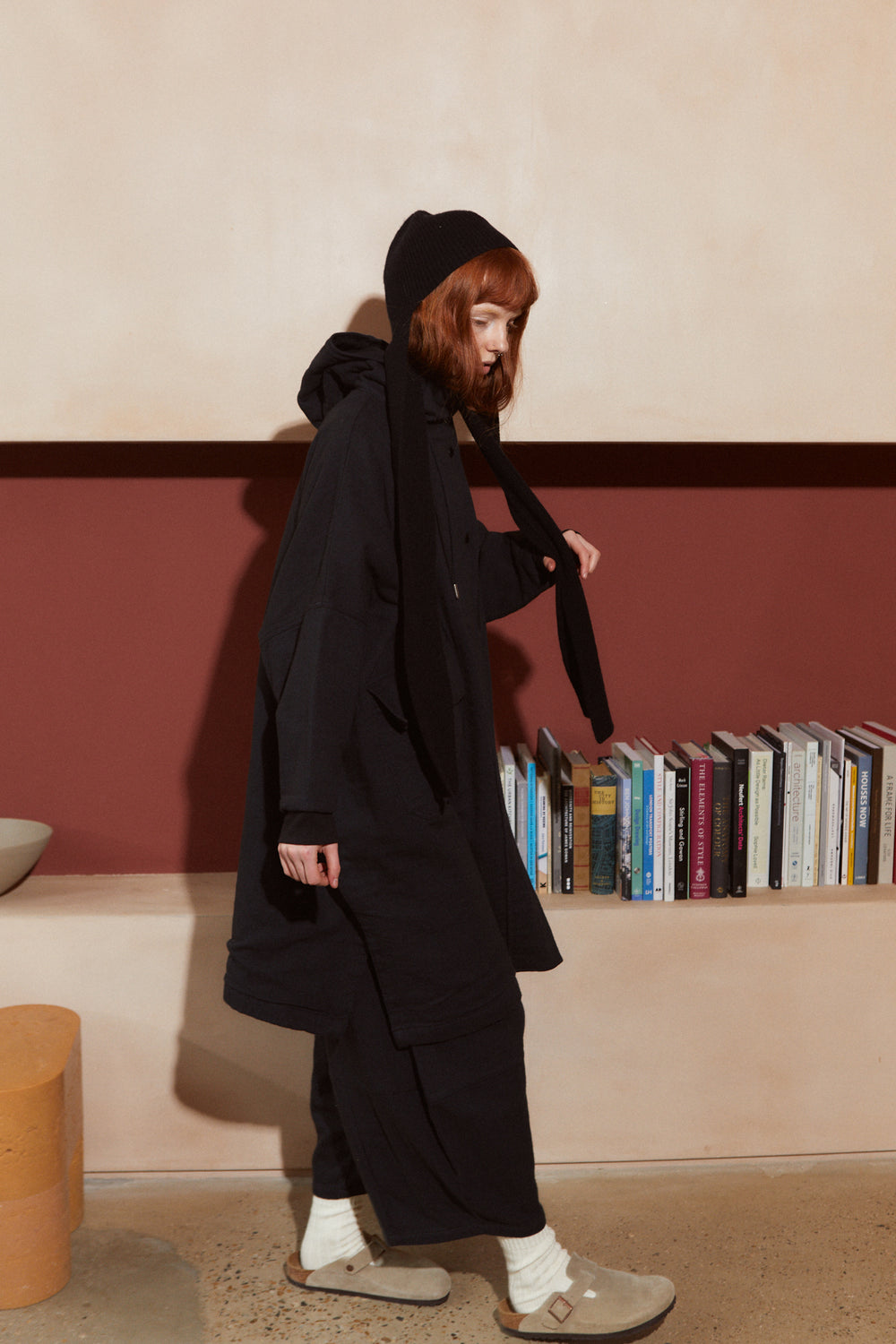 A model with red hair wearing a L.F.Markey Rib Scarf Beanie Black, long black coat, black pants, white socks, and beige slip-on shoes, standing in a room with a bookshelf in the background.