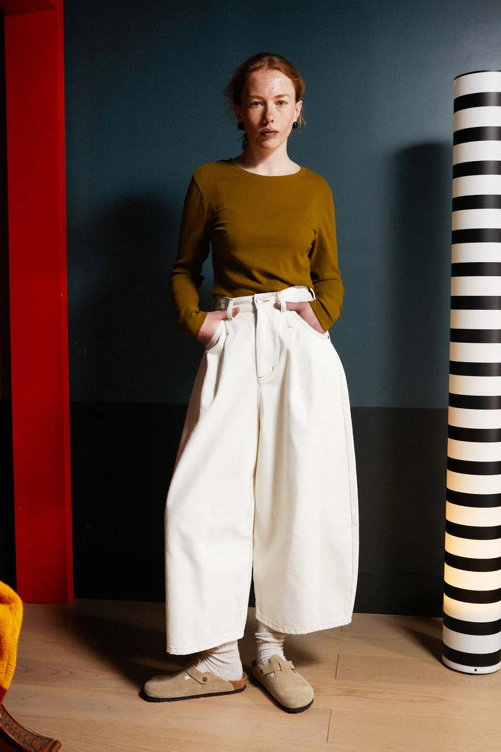 Person wearing a mustard top and white pants standing in a room with a striped column.