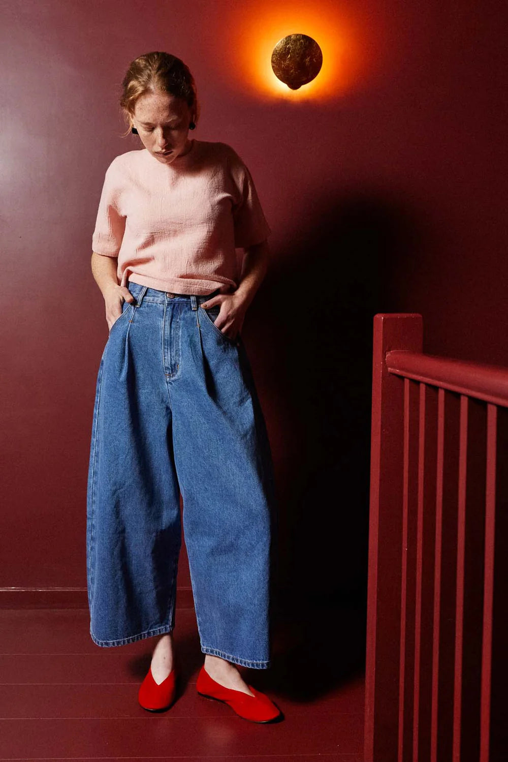 Person wearing a pink top and blue jeans standing against a red wall with a light fixture.