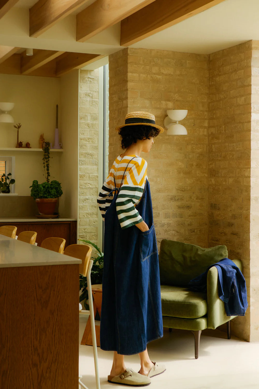 Woman in a striped sweater and a LF Markey denim blue dress standing in a modern interior space with wooden beams and a green chair.