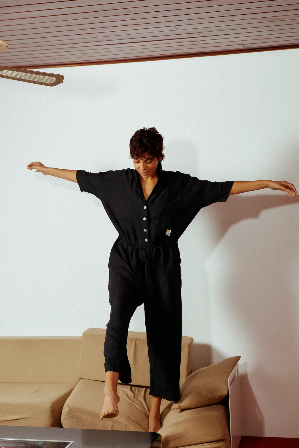 Person wearing a black jumpsuit standing in a room with a beige couch and white walls.