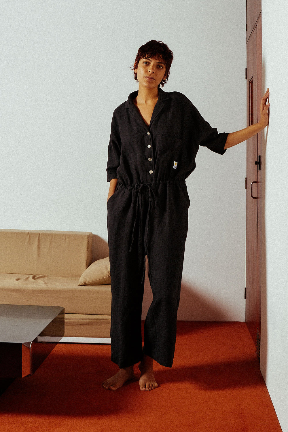 Woman in a black jumpsuit standing in a room with a beige sofa and red carpet.