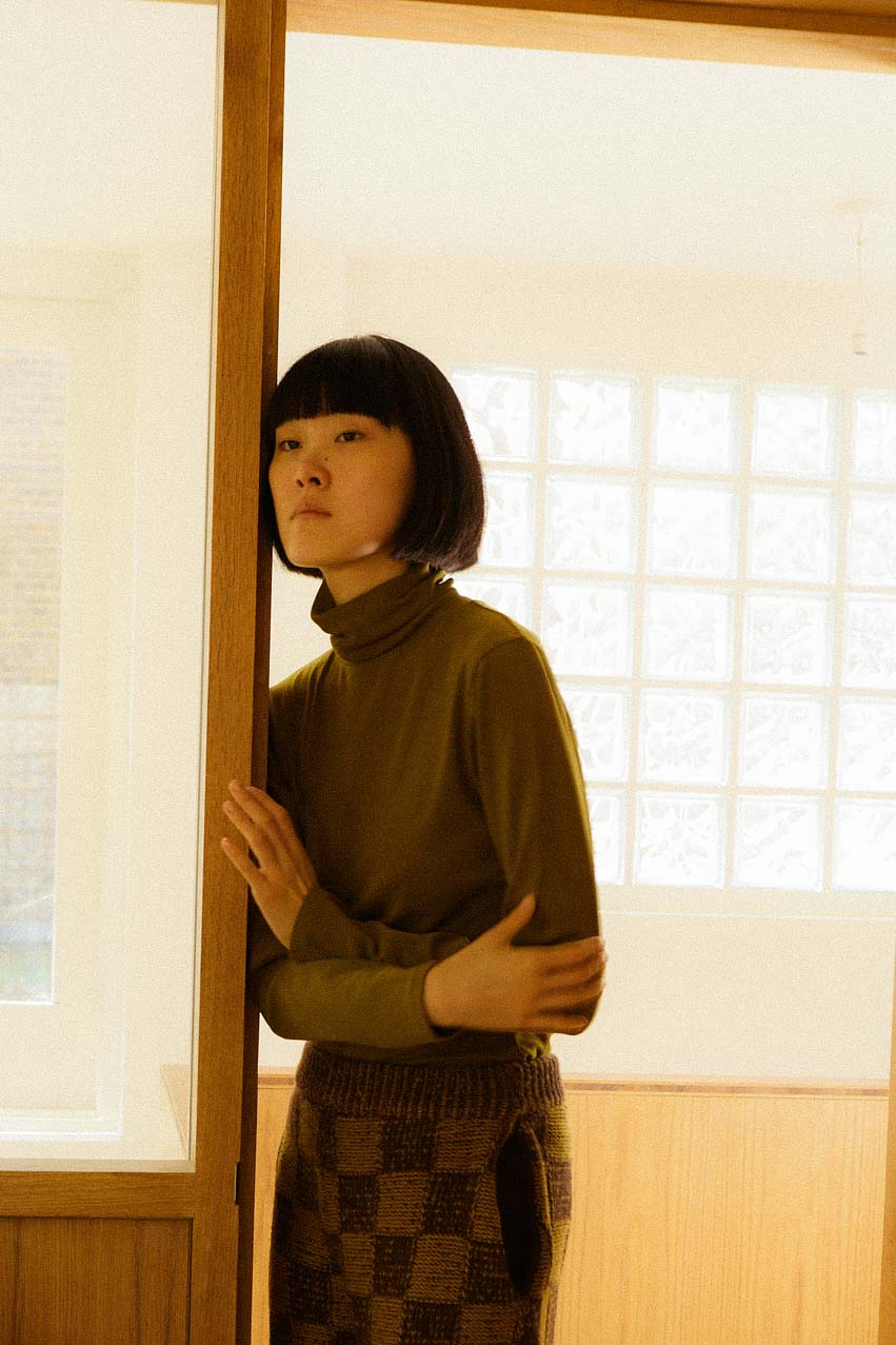 Person wearing a avocado LF Markey turtleneck top and checkered skirt standing in a room with light-colored walls and a window.