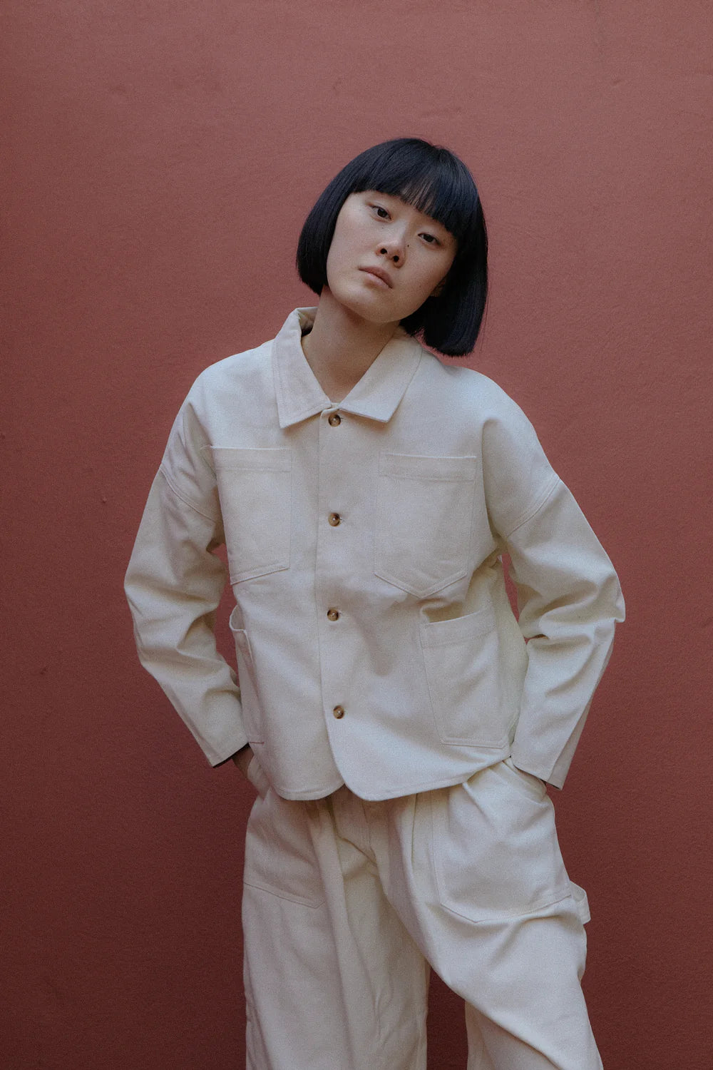 Person wearing a beige jacket and trousers against a red-brown wall