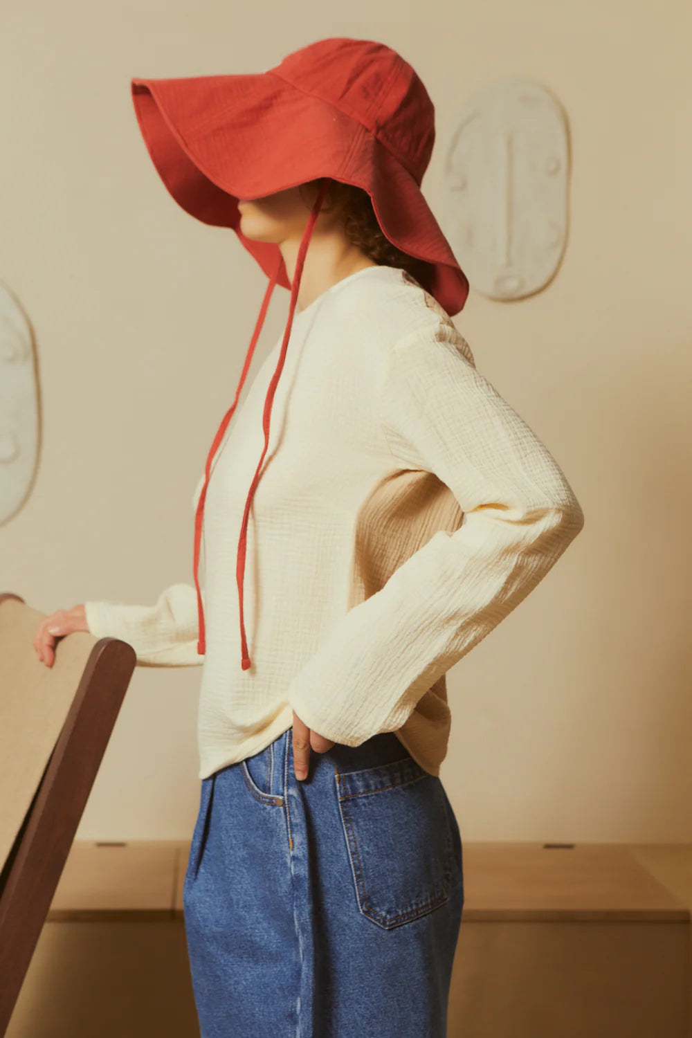 Person wearing a soft red sun hat, white sweater, and blue jeans in a neutral setting