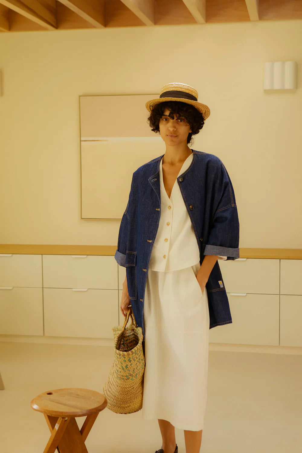Person wearing a blue jacket and ecru top and skirt with a straw hat, standing in a room with wooden ceiling and white walls.