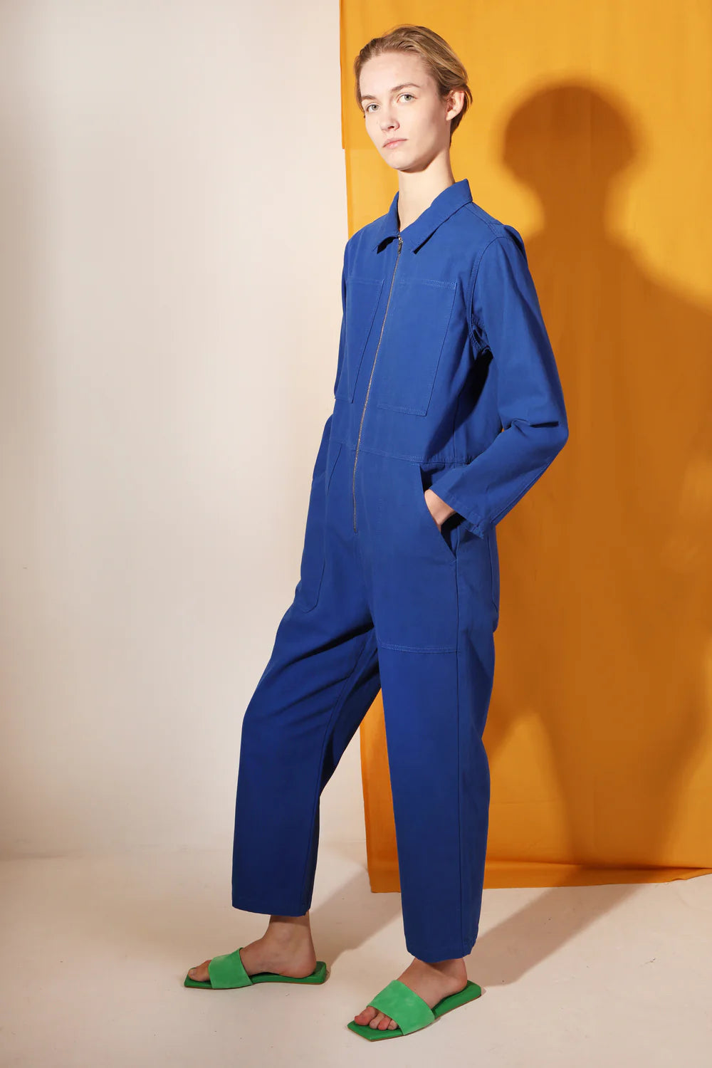 Person wearing a blue LF Markey jumpsuit standing against an orange curtain.