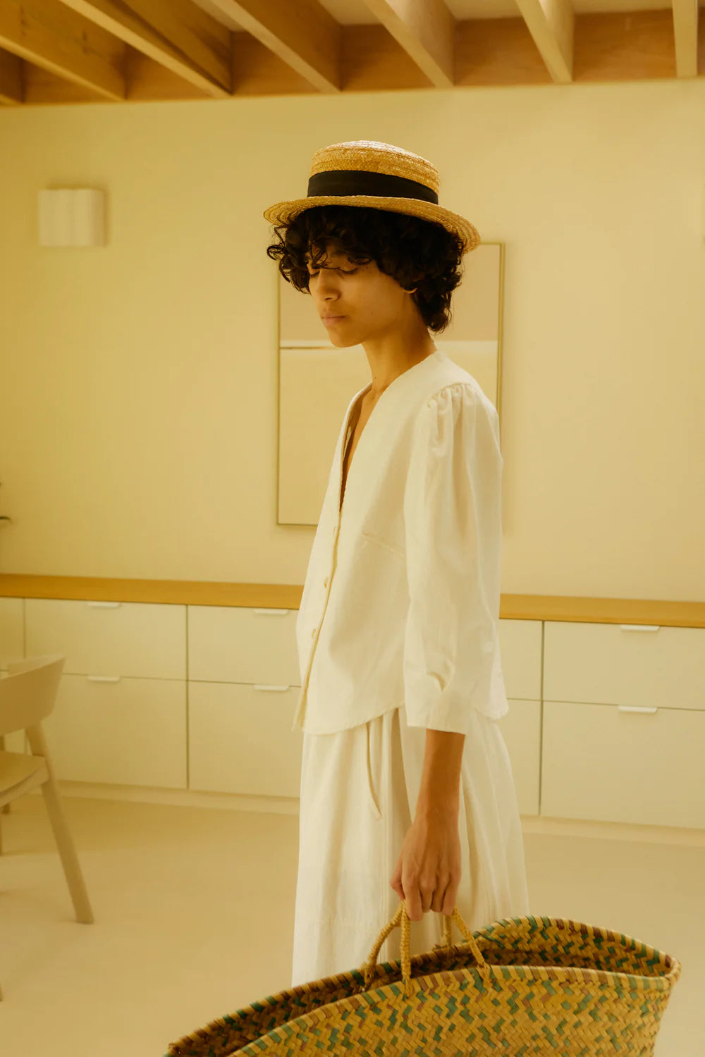 Woman in a ecru top and skirt holding a woven bag in a minimalistic room.